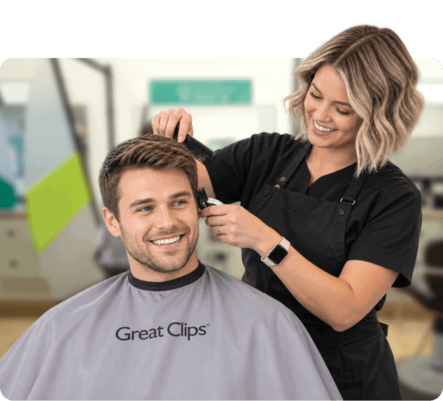 Smiling Great Clips hairstylist uses clippers and a comb to trim a male customer's hair as he sits in a salon chair wearing a branded cape.