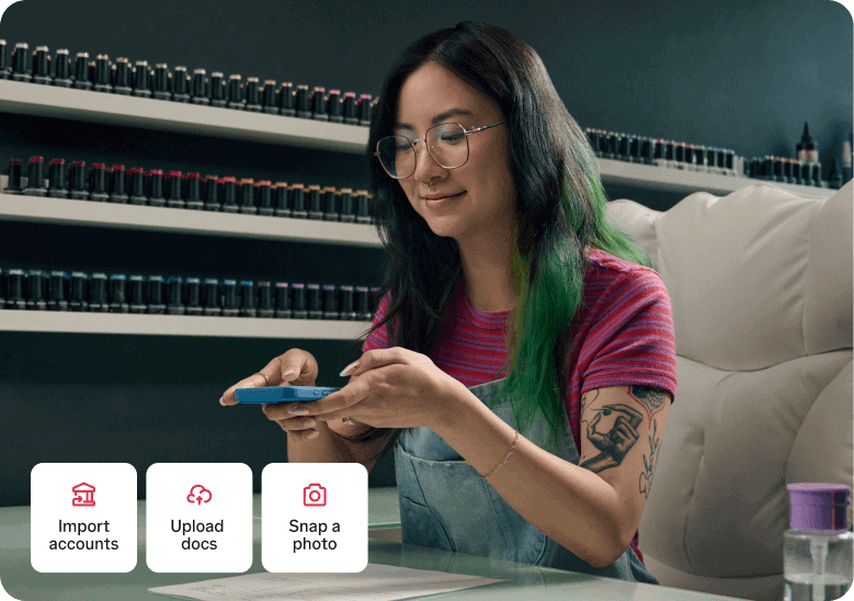 A woman with glasses and green-tipped hair uses her smartphone in a nail salon, with TurboTax options for importing accounts, uploading documents, and snapping a photo displayed below.