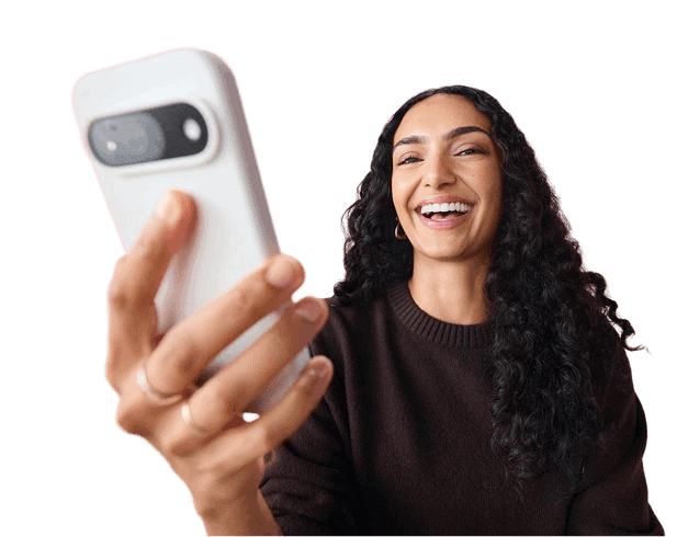 A smiling woman with long, curly dark hair holds up a white smartphone while wearing a dark sweater, isolated on a transparent background.