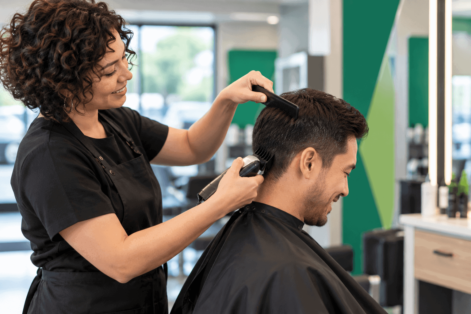 Smiling hairstylist uses clippers and a comb to trim a male customer's hair in a bright, modern salon with green accents.