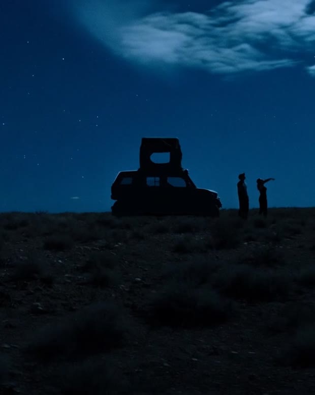 Two people stand beside an overland vehicle with a rooftop tent, silhouetted against a starry desert sky at night.