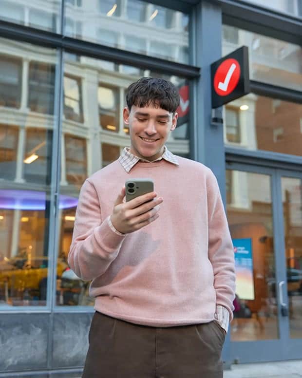 A young man in a pink sweater smiles at his phone outside a glass storefront with a TurboTax checkmark sign.
